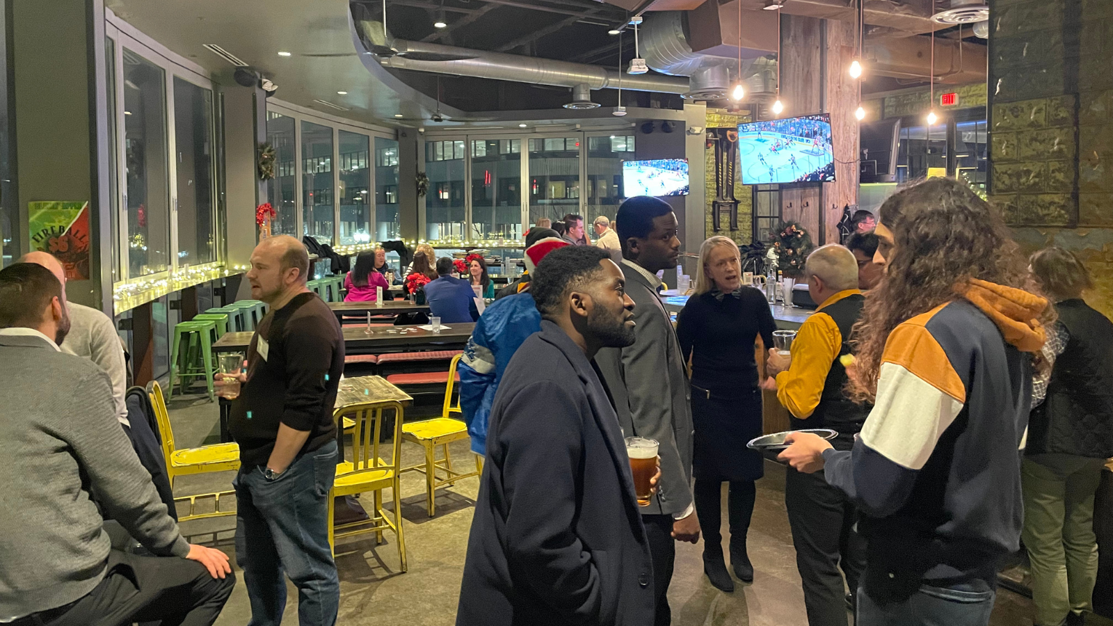 A photo of a lively group of software professionals standing and talking in a modern bar/restaurant setting with large windows overlooking a city at night. Attendees are dressed business-casual and holding drinks, engaging in conversation. The venue has exposed ceiling ductwork and industrial lighting.