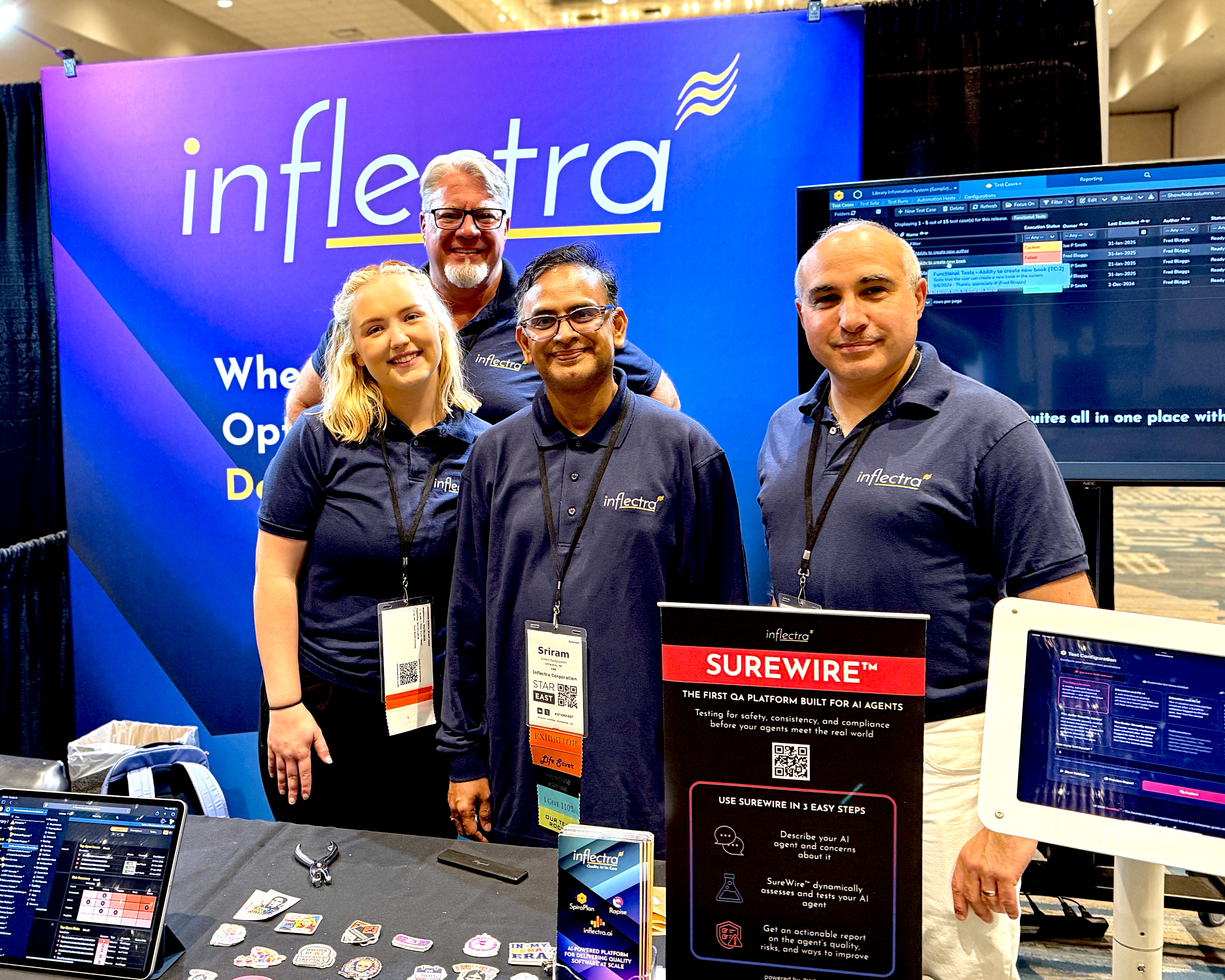 Four members of the Inflectra team stand smiling in front of a bright blue and purple exhibit booth at STAREAST. They are all wearing navy blue Inflectra polo shirts and conference badges. In the foreground, a tabletop display features a sign for SureWire, described as the first QA platform built for AI agents, alongside a collection of colorful laptop stickers and a tablet showing a software dashboard. A large monitor in the background displays a technical test management interface.